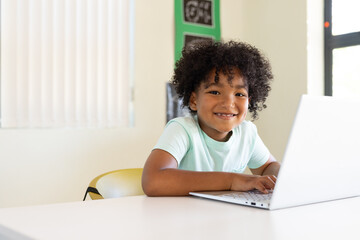 African American schoolboy sitting at white table in classroom typing on silver laptop in mint tee