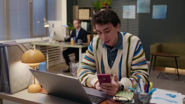 Rack focus of adult Caucasian man interrupting young male colleague video calling on smartphone instead of working on laptop in shared office workspace