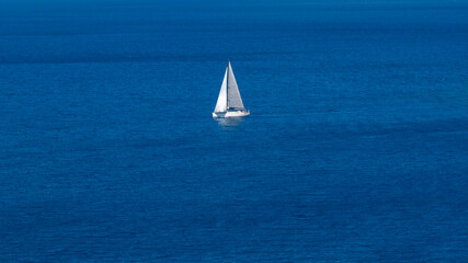 Fototapeta premium High-angle shot of a white sailboat with its sails up, navigating through a vast and tranquil deep blue sea. The minimalist composition emphasizes the scale and serenity of the ocean.