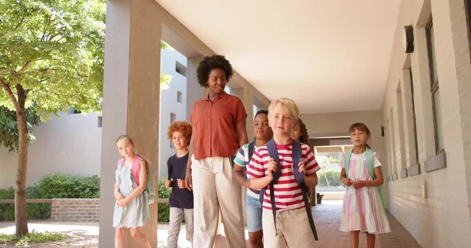 Teacher with children guiding along walkway with columns backpacks as blonde boy stepping to lead