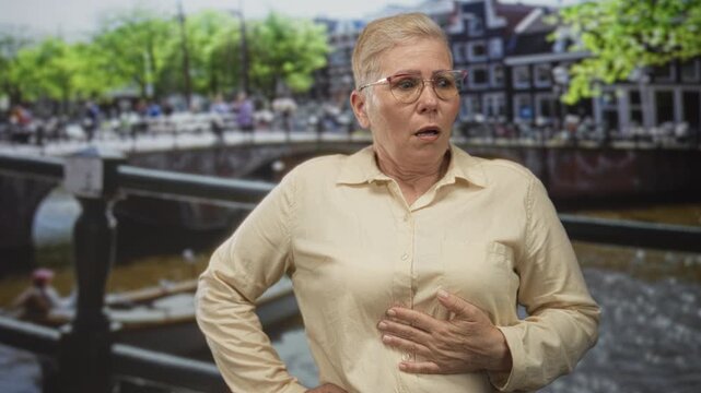 Middle age woman clutch chest for heartattack on street bridge in amsterdam by canal and boats wearing glasses and beige shirt; alarm health concern.