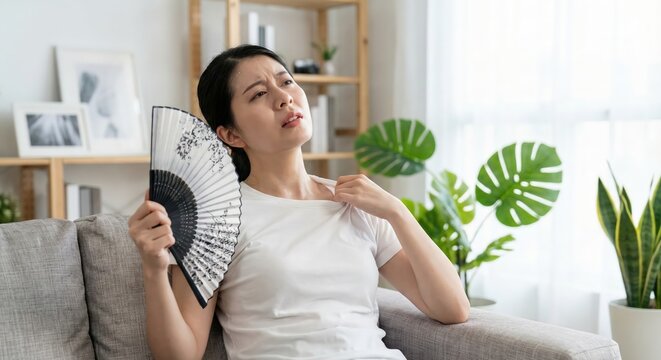 Distressed Young East Asian Woman Sitting on Sofa in Bright Living Room Fanning Herself with Handheld Fan During Summer Heatwave, Uncomfortable Woman Feeling Hot Indoors in White T-shirt