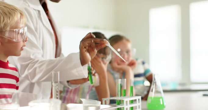 Male instructor in labcoat teaching kids with goggles dipping pipet into flask filling tubes in lab