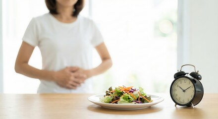 Naklejka premium Hungry young adult woman in white t-shirt holding stomach while waiting for intermittent fasting time to end near fresh salad and alarm clock indoors, healthy lifestyle and weight loss concept.