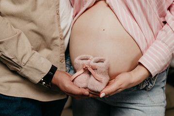 A pregnant girl shows off her belly, her beloved man hugs her, they hold pink shoes of the future baby woven from threads in their hands during a photo shoot