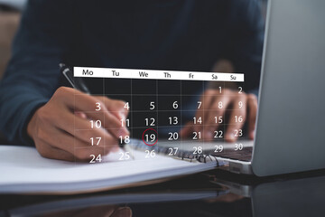 Person actively planning and scheduling tasks, marking an important date on a digital calendar overlay while writing notes in a notebook next to a laptop in a focused workspace.