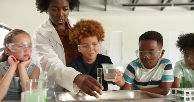Lab teacher pointing to cylinder, guiding kids pouring cup into tray in lab revealing green color