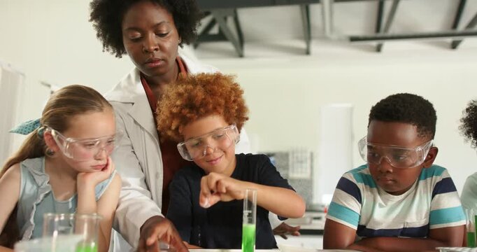 Teacher guiding 3 children placing tablet into cylinder in lab wearing goggles observing reaction