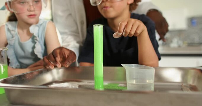 Teens in goggles at lab, teacher guiding student adding tablet into cylinder, forming green foam