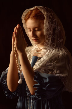Caucasian teenage girl praying with closed eyes, holding rosary beads in hands, wearing lace headscarf and vintage style dress, standing against dark background