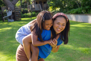Fototapeta premium Mother and daughter giving piggyback ride on green lawn in backyard next to wooden treehouse