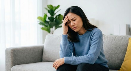 Obraz premium Southeast Asian woman sitting on a grey sofa in a bright living room, holding her head with a pained expression suffering from a migraine or stress, wearing a blue sweatshirt, mental health concept