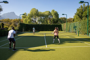 Fototapeta premium Diverse friends playing doubles tennis with rackets by net and hedges on backyard court