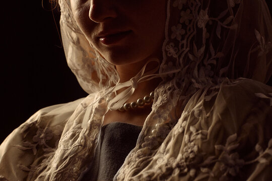 Caucasian young adult woman wearing lace headscarf and pearl necklace standing in low light, face partially visible, lips and chin softly illuminated, traditional style clothing