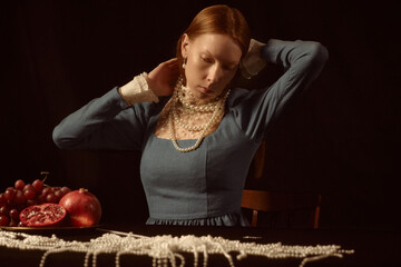 Caucasian young adult woman adjusting pearl necklace while sitting at table with grapes, pomegranate and strands of pearls, wearing vintage style dress, looking downward