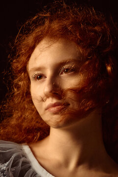 Portrait of Caucasian young adult woman with curly red hair looking slightly away from camera, soft facial expression, closeup of face and shoulders, subtle wind blowing hair