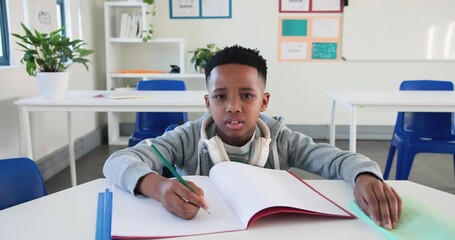 African American boy writing with green pencil in notebook at school table pausing counting fingers - Powered by Adobe