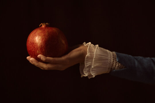 Young adult woman hand holding pomegranate against dark background, delicate lace cuff visible, fruit resting gently on open palm, classic composition