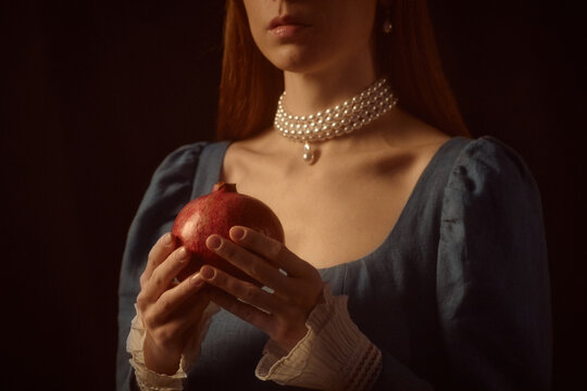 Caucasian young adult woman holding pomegranate with both hands, wearing pearl necklace and vintage style dress, standing against dark background, face partially visible