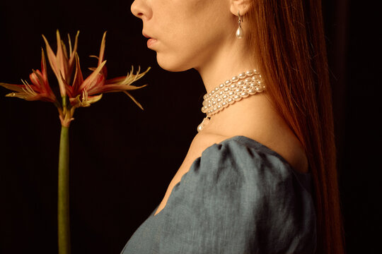 Caucasian young adult woman standing in profile holding exotic flower, wearing pearl necklace and earring, long straight hair visible, neutral expression, dark background