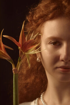 Portrait of Caucasian teenage girl with curly red hair looking into camera, holding orange lily flower near face, showing delicate features and subtle jewelry against dark background