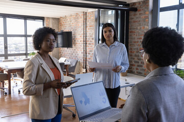 Diverse female coworkers reviewing laptop, tablet, printed documents at modern open-plan office