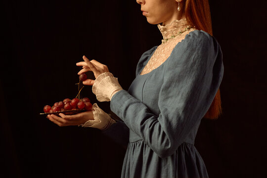 Young adult Caucasian woman holding plate of red grapes, gently picking fruit with delicate fingers, wearing vintage style dress, standing against dark background