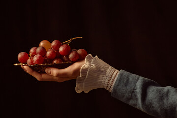 Woman holding plate of red grapes with delicate lace cuff visible against dark background, hand extended forward presenting fruit in classic art style