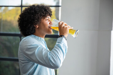 Adult african american man in blue shirt standing by window drinking orange juice from glass