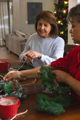 Middle-aged and senior female friends crafting wreaths on wooden table in living room with mugs
