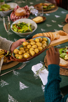 Diverse friends passing oval dish of boiled potatoes on dining table at home with breadboard