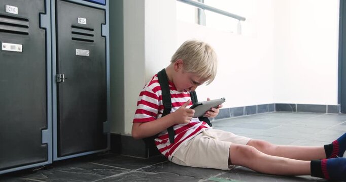 Schoolboy sitting on tiled corridor floor holding tablet adjusting straps waiting between classes