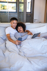 Fototapeta premium Diverse male couple reclining on bed in bedroom with gray duvet holding smartphone and tablet