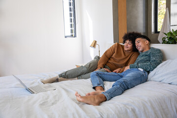 Two men reclining together on bed in bright bedroom beside silver laptop with gold lamp