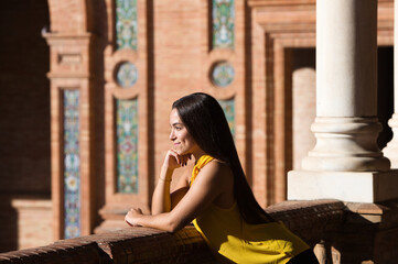 Fototapeta premium A young, beautiful, dark-haired Latin woman wearing a yellow shirt and black trousers leans on the railing of the Plaza de España with her chin resting on her hand, looking attentive and happy.