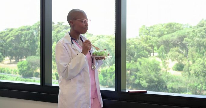 Adult African woman on break eating salad with wooden fork at window, lab coat, copy space