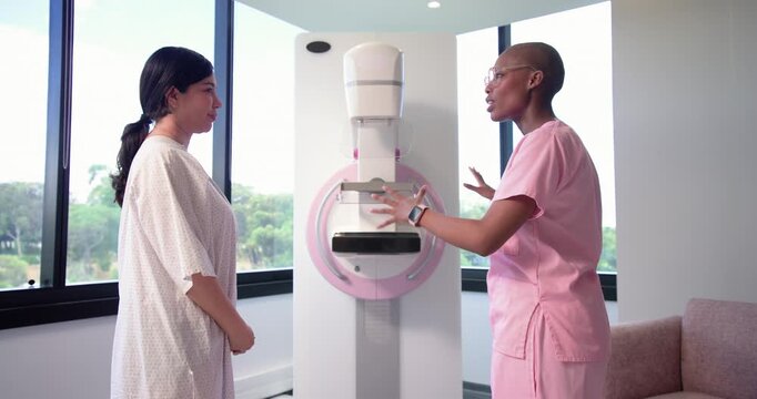 Female tech guiding patient in gown in clinic after explaining, aligning breast on paddle for scan