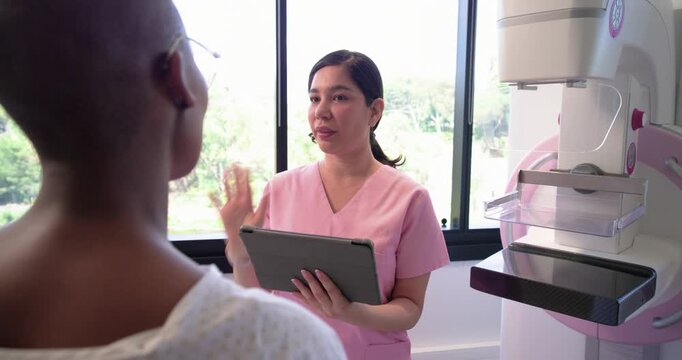 Female tech explaining mammogram to female patient in imaging room using tablet pointing to machine