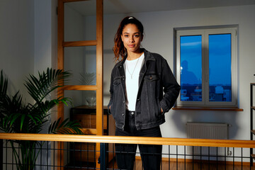 Portrait of young adult biracial woman standing indoors with hands in pockets looking into camera, confident expression, visible window and plant in background evening setting