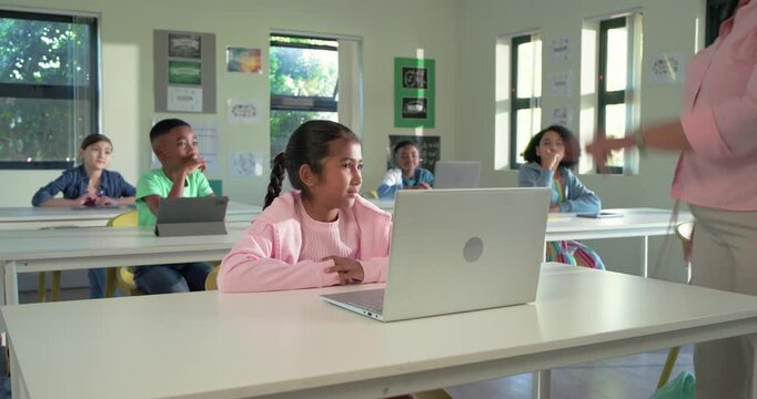 Female child in pink top turning to teacher entering from right in classroom, listening at laptop