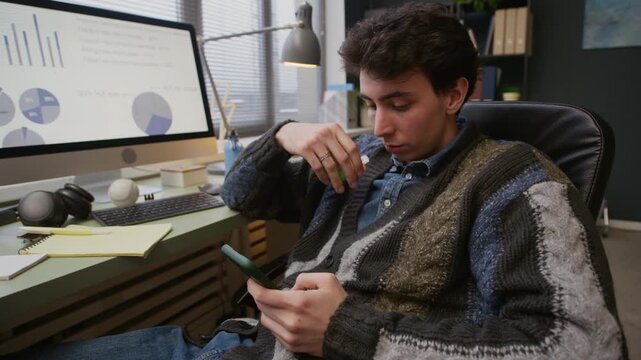 Zoom out shot of young Caucasian man smoking vape and using smartphone while sitting at office desk in front of computer monitor with diagrams on screen