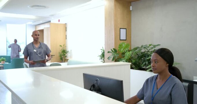 African American male doctor in scrubs entering left, using tablet at front desk checking records