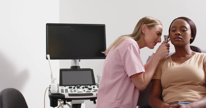 Female nurse in pink scrubs doing otoscope exam as African American patient angling head in clinic