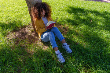 Fototapeta premium Young woman with curly hair reading a book under a tree in a park enjoying outdoor leisure and learning