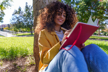 Young woman concentrating, writing notes in red notebook while sitting by a tree in a city park, enjoying nature