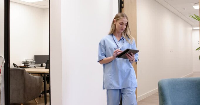 Female nurse in scrubs with stethoscope entering corridor, tapping tablet, reviewing chart