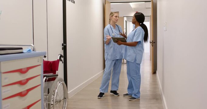 Diverse female medical staff in scrubs reading tablet on patient update in hall, pointing for care