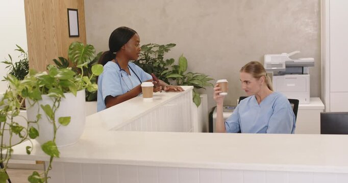 Diverse female nurses in scrubs on break talking, leaning at reception, sipping paper cups, smiling