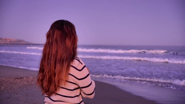 A woman with long red hair stands on a beach looking out at the ocean. The sky is a beautiful purple color, and the water is calm. The scene is peaceful and serene
