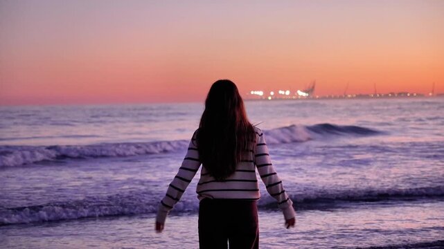 A woman stands on the beach looking out at the ocean. The sky is a beautiful mix of pink and purple hues, creating a serene and peaceful atmosphere. The woman is lost in thought